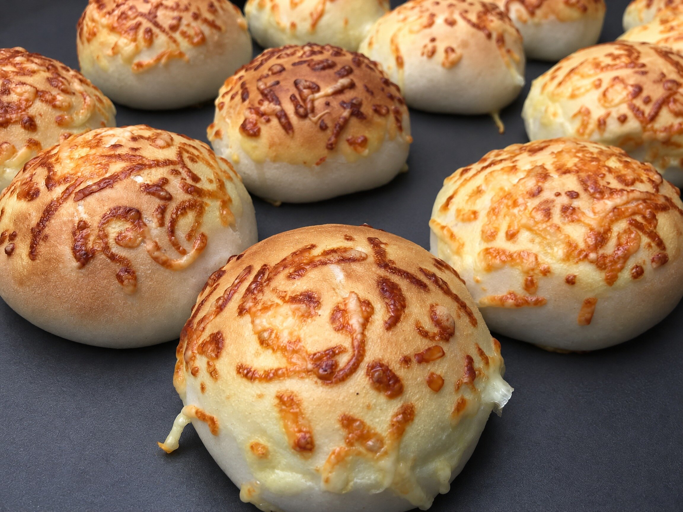Baked buns with a pattern of melted cheese on top, on a dark baking tray
