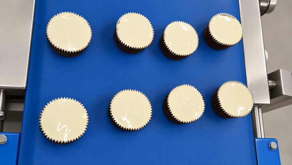 Top view of eight muffins in dark paper cups on a blue conveyor, each covered edge to edge with a white topping